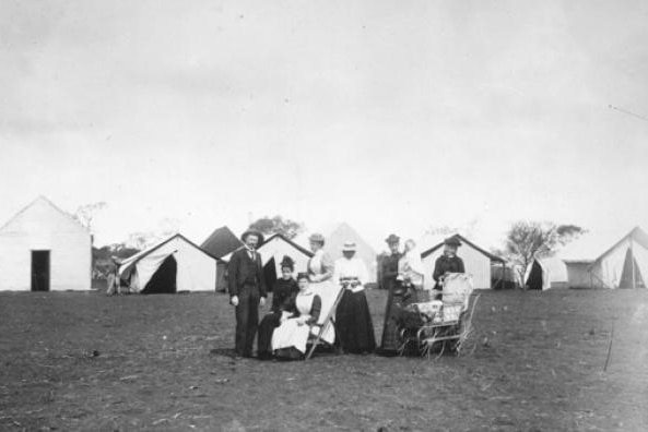 An old black and white photo of women in long dresses and hats and men in suits in front of a makeshift tent hospital