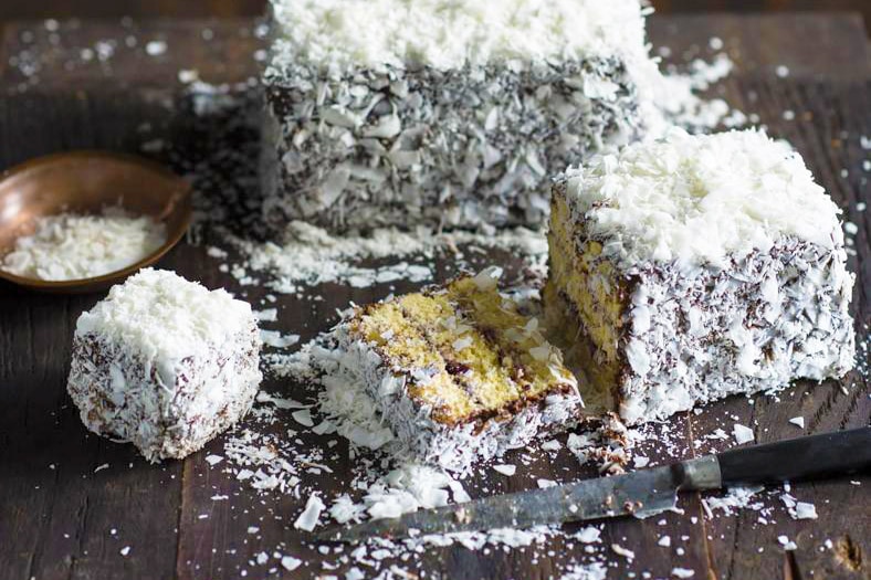 Sliced lamington cake covered in lots of coconut on a timber table for a story about lamington's history and baking tips.