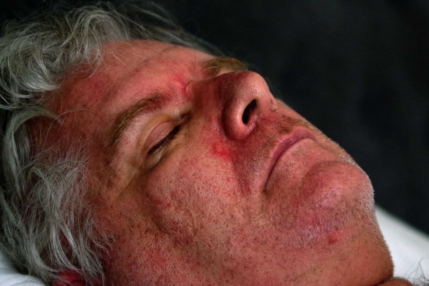 A close-up shot of a man with grey hair lying down on his back with a hand on his chest.