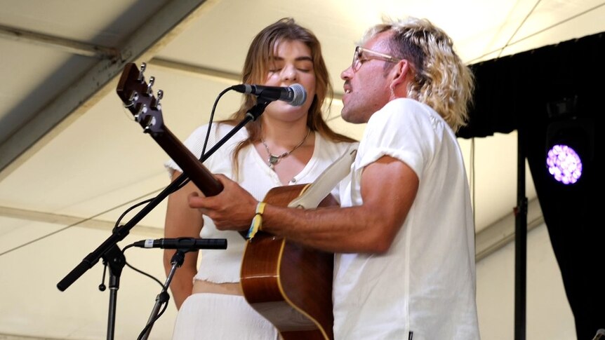 A man with a unique hair cut sings and plays guitar on stage with a young woman