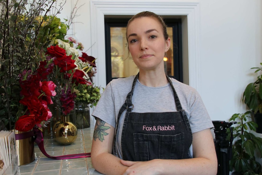 A woman stands at a counter in a flower shop posing for a photo in front of bouquets of flowers on her right.