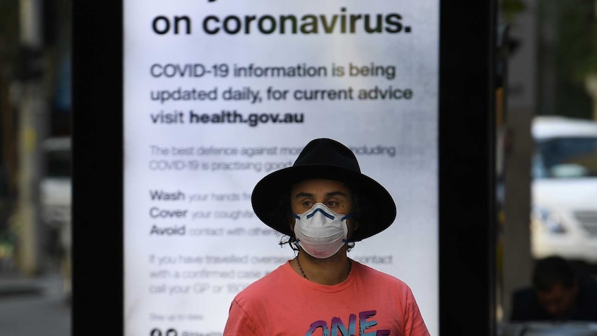 A man in a hat and a face mask stands in front of a sign that reads "state informed on coronavirus".