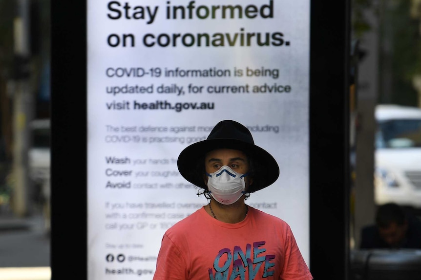 A man in a hat and a face mask stands in front of a sign that reads "state informed on coronavirus".
