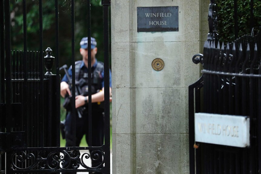 A man holding a gun wearing a police vest stands at a stately gate.