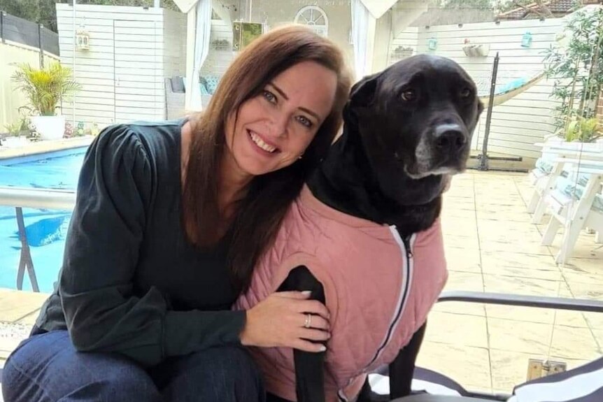 a woman smiling while learning up against her dog, which is in a vest