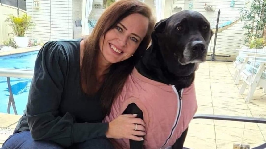 a woman smiling while learning up against her dog, which is in a vest