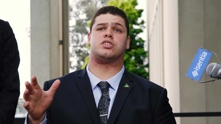 Young man wears dark suit jacket, pale shirt and dark tie while conducting media interview