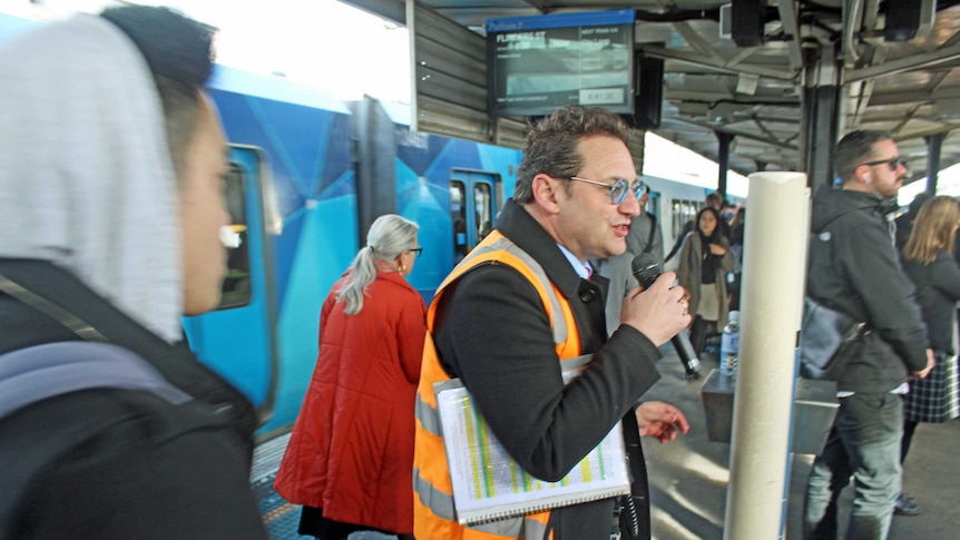 A man in a high-vis vest stands on a train platform talking into a microphone, as commuters walk around him and a train waits,