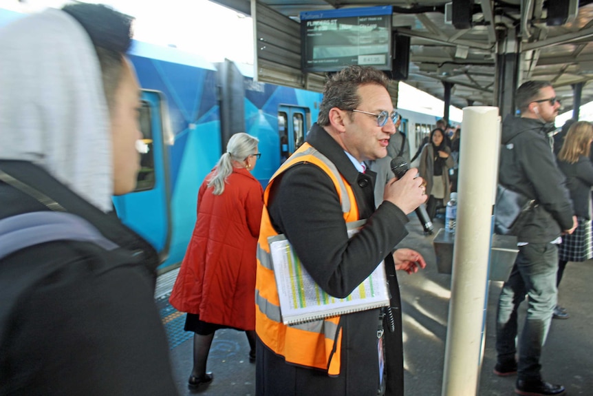 A man in a high-vis vest stands on a train platform talking into a microphone, as commuters walk around him and a train waits,