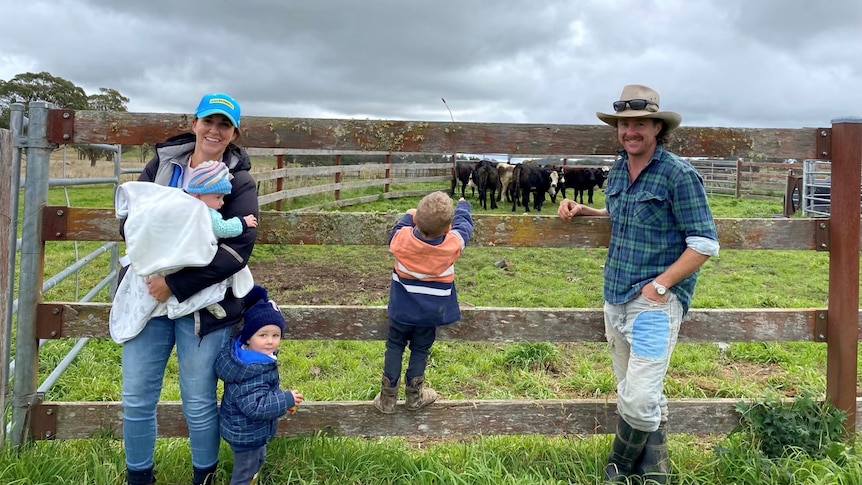 Alison Jones standing by a cattle pen holding her third baby, with her other two children next to her and husband on the right 