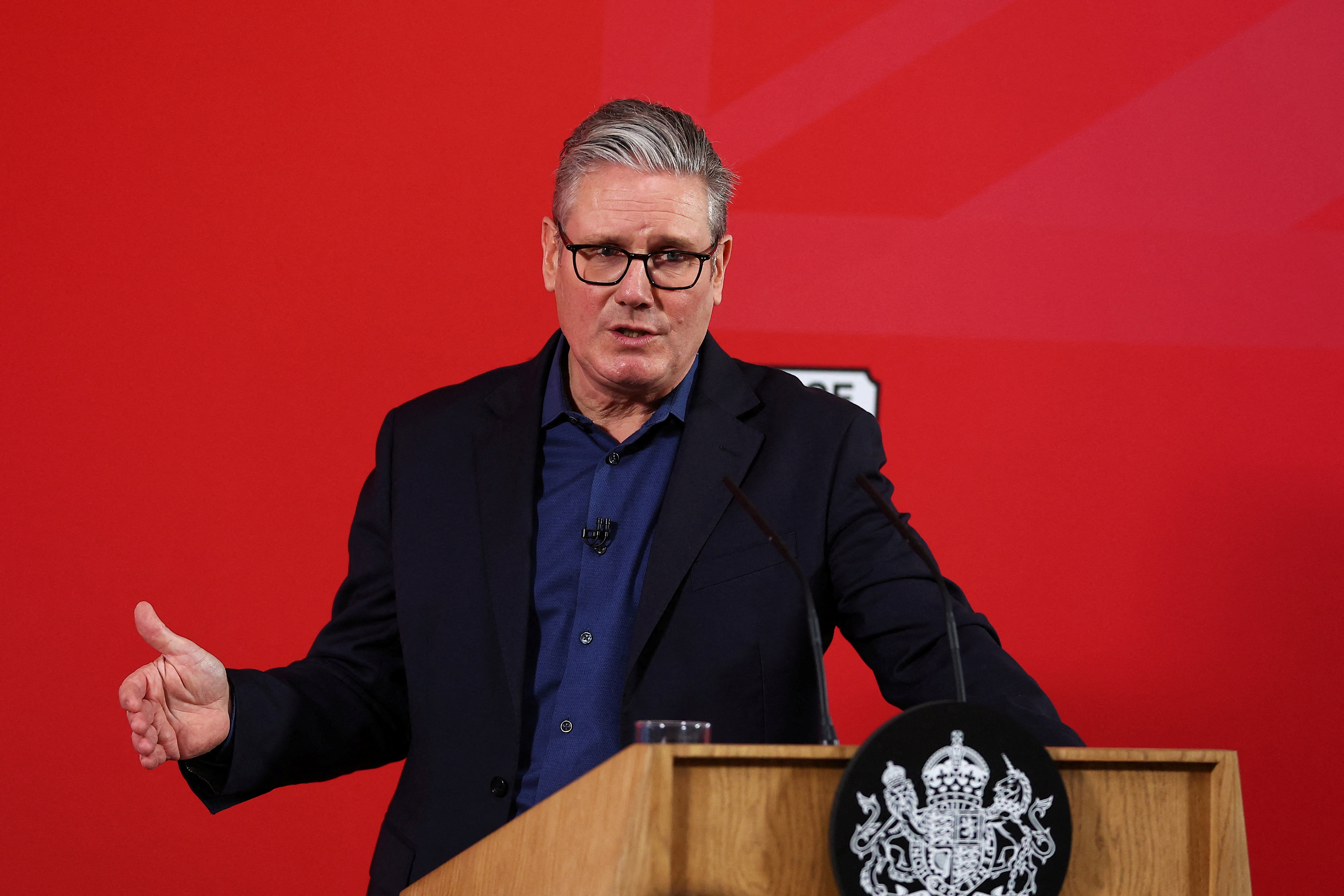 Keir Starmer, sin corbata, gesticula mientras habla sobre un fondo rojo brillante.