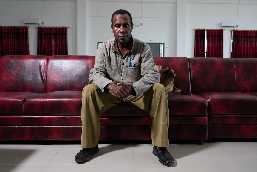 Silas Kalami, appearing sombre, sits on a large couch, his hands clasped in front of him.