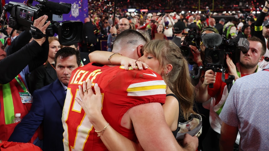 A woman hugs a man, her eyes are closed. They're surrounded by people but look peaceful