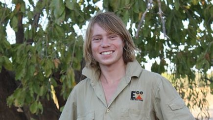 Josh, looking tanned and wearing a khaki collared shirt, smiles at the camera, in front of a tree.