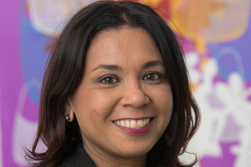 A well dressed woman with dark hair stands in front of pink, purple, white and orange coloured Aboriginal art.