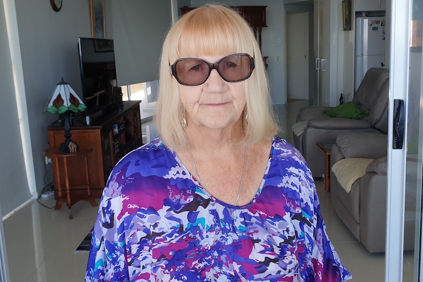 A woman with a blonde bob wearing sunglasses and a floral top looks towards the camera.