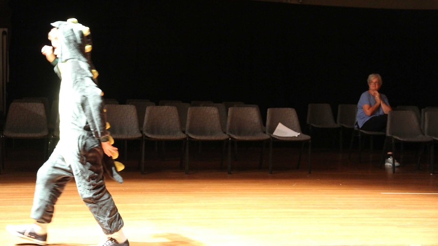 Boy in costume on the stage floor whilst director watches from the sidelines