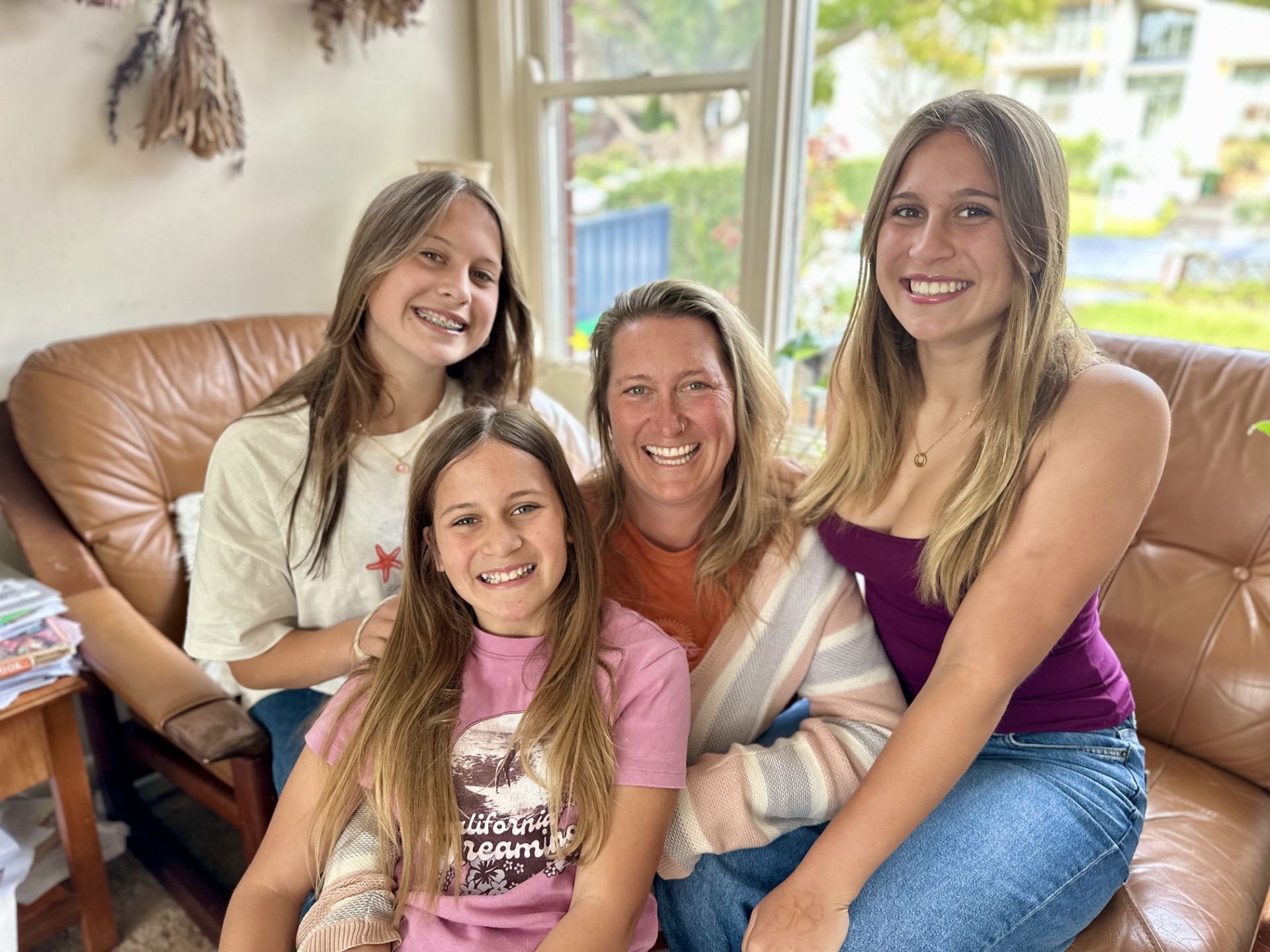 Three smiling teenaged girls sit cosily around their mum in a loungeroom.