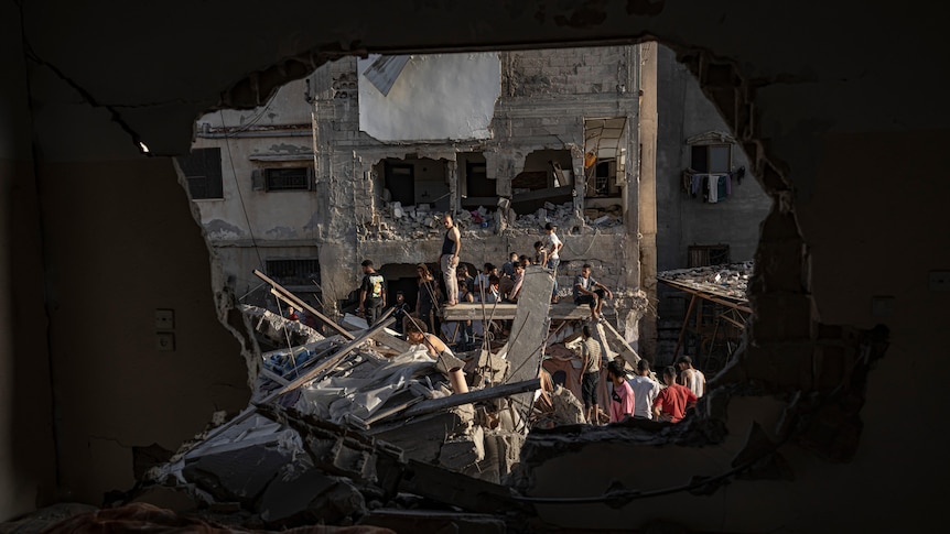 through a hole blasted into a building's wall, neighbouring buildings have also been reduced to rubble, people standing on them