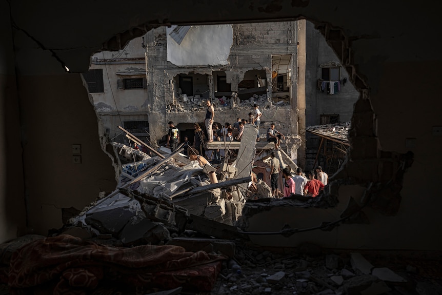 through a hole blasted into a building's wall, neighbouring buildings have also been reduced to rubble, people standing on them