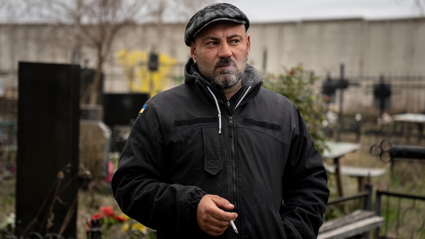 A man in a heavy black jacket with a Ukrainian flag on the shoulder, stands smoking in a graveyard on an overcast day