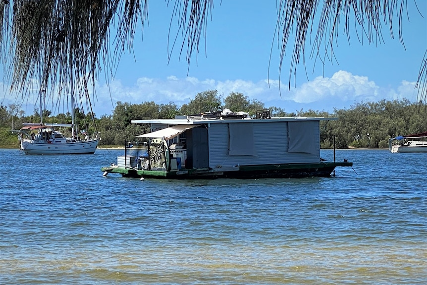 Grey house boat floating in water with covers over windows, a BBQ on back deck and a generator and other objects on roof. 