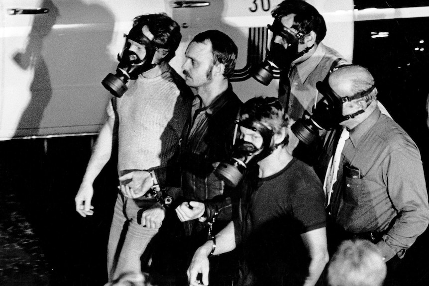 A man walks down a street in cuffs, surrounded by men in gas masks 