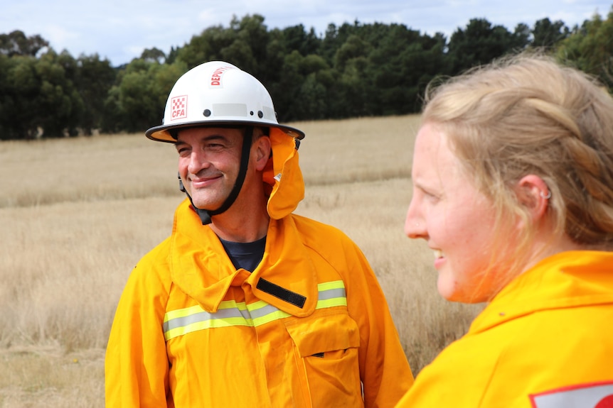 A man dressed in firefighting gear including helmet smiles with a woman looking on.