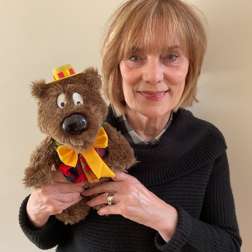 woman with shoulder-lend sandy hair with a fringe holding a plush bear toy wearing a waistcoat