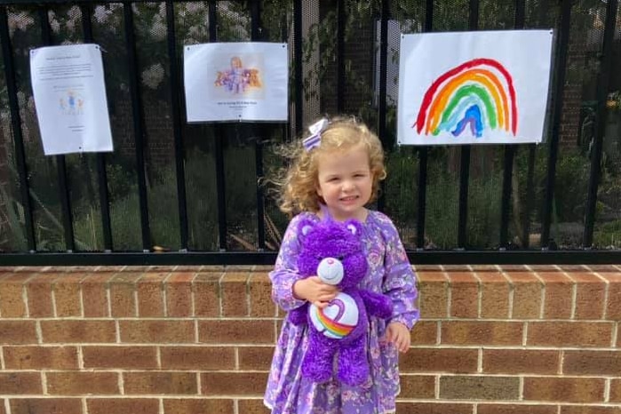 A young girl in a purple dress stands in front of a fence that has a picture of a rainbow stuck to it.