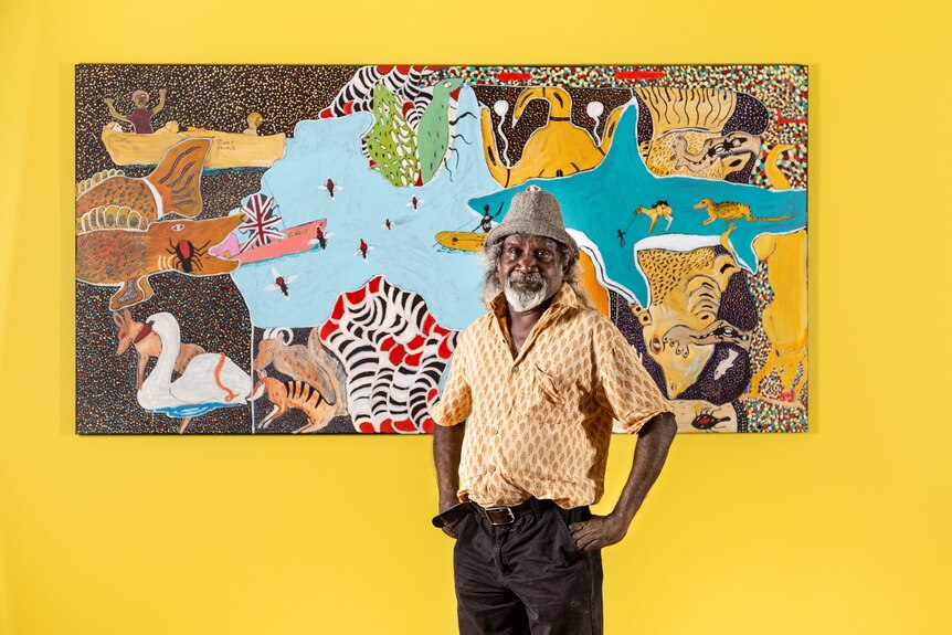 An older Aboriginal man in hat and shirt stands in front of a colourful painting on a yellow wall