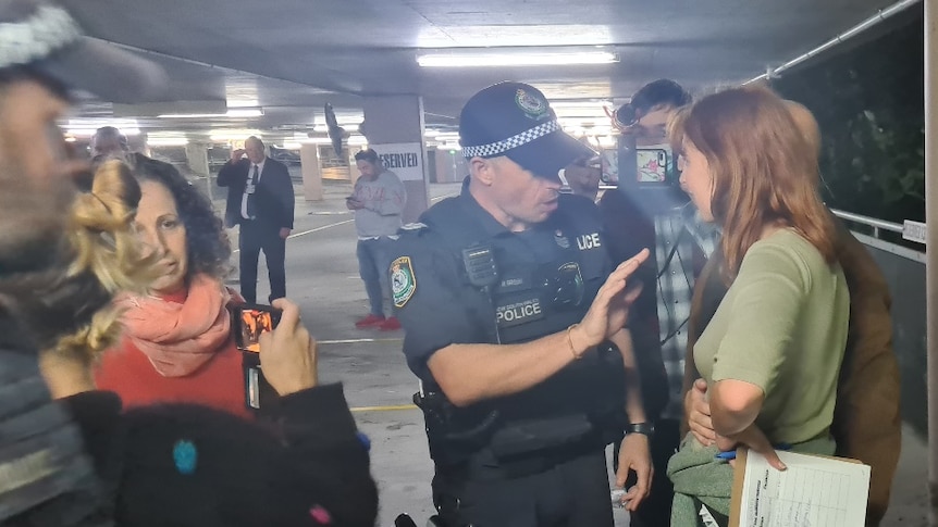 A police officer gestures as he speaks with a woman in a covered car park as other police and people mill around.