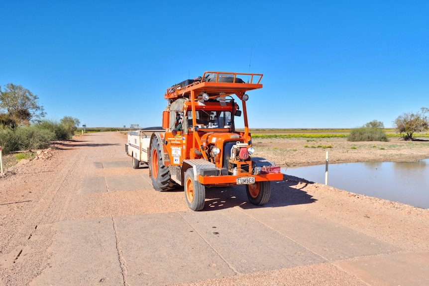 An orange tractor on a dirt road in the outback with a blue sky behind it towing a trailer.