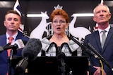 Blank-faced, James Ashby and Steve Dickson flank Pauline Hanson as she speaks at a lectern with microphones in front.