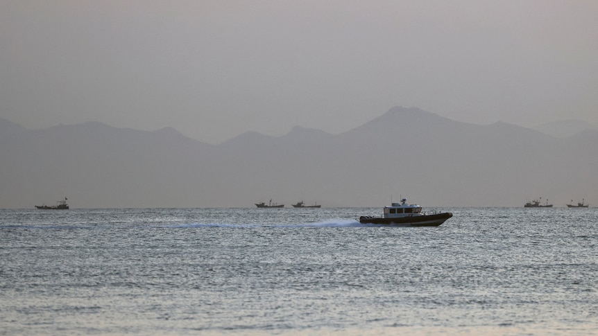 A coastguard motors along the sea with boats and mountains in the background