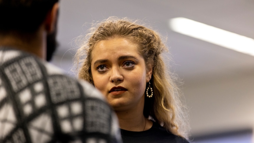Young woman with curly blonde hair, dark eyebrows and brown eyes wears black shirt and white beaded earrings and looks doubtful