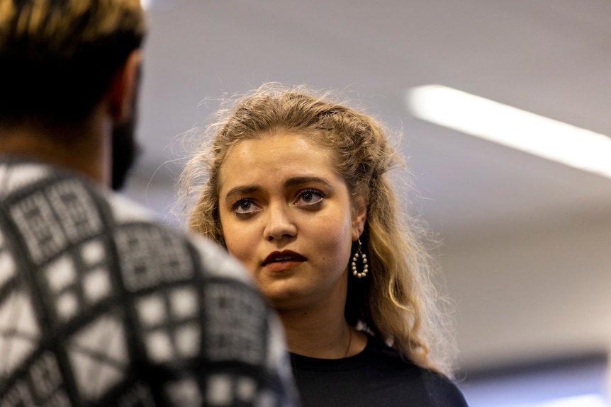 Young woman with curly blonde hair, dark eyebrows and brown eyes wears black shirt and white beaded earrings and looks doubtful