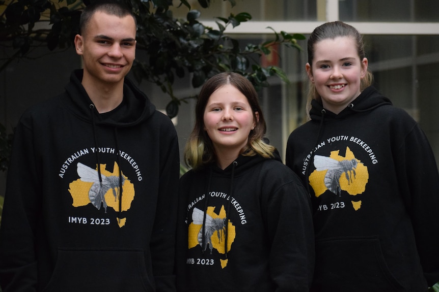 Three teens stand smiling, wearing black hooded jumpers with a bee on the front and Australia