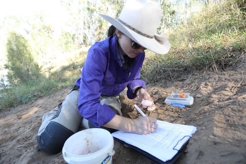 A woman in a purple shirt and widebrim hat, kneels on a river bank writing on a notepad on the ground with an egg in one hand