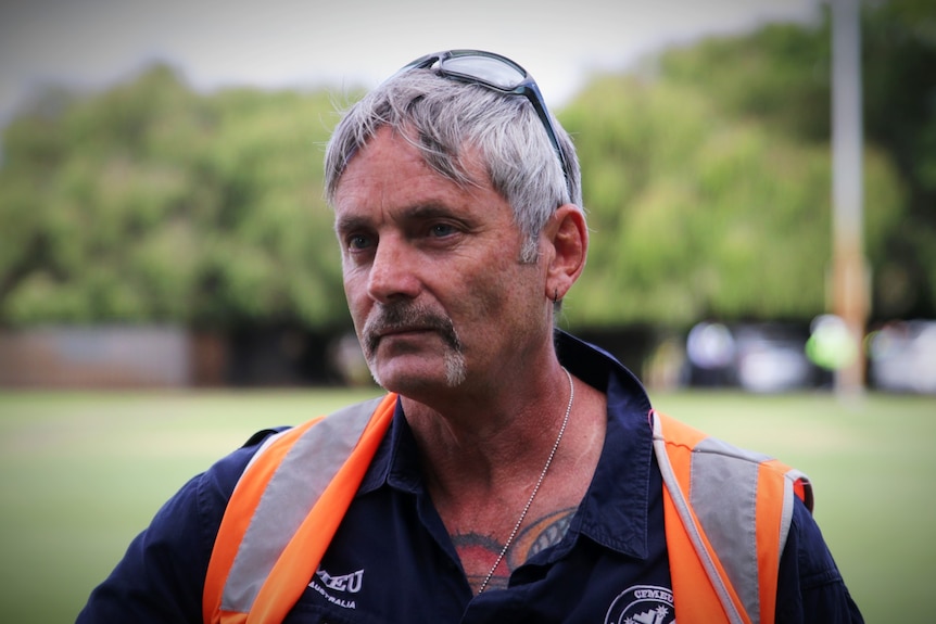 A man wearing a high-vis jacket and a blue collared shirt, with chest tattoos, stands in a park.