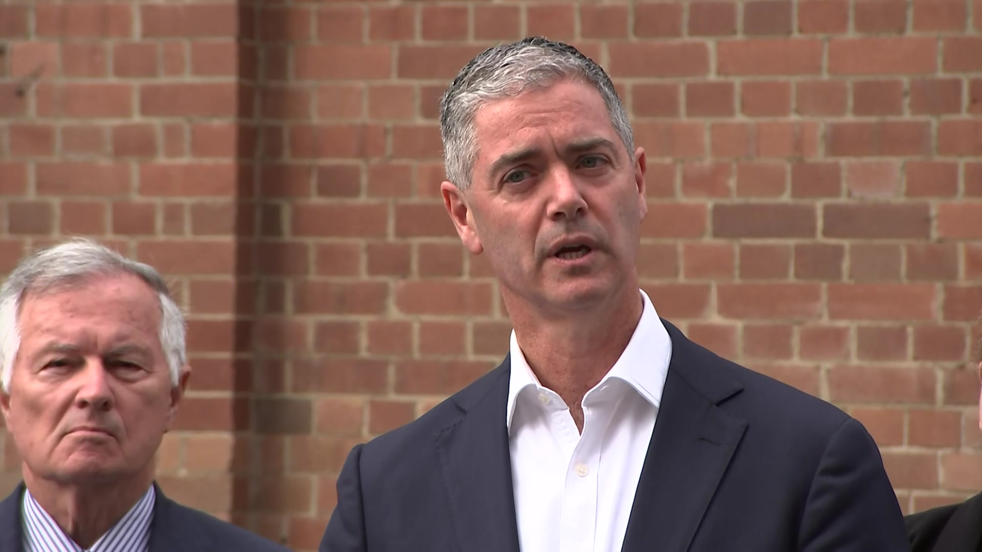 Art Minister John Graham in a white shirt, black blazer standing in front of a brick wall