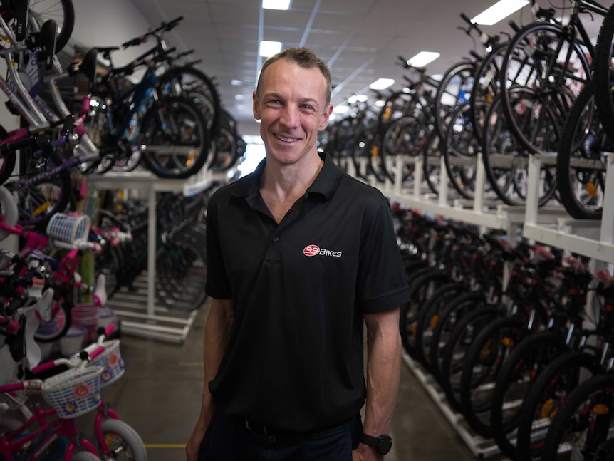 A man wearing a black polo shirt stands in shop surrounded by bicycles.