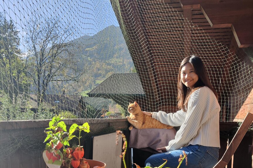 A woman in a white jumper and jeans sits on a deck in the sun, patting an orange cat. In the background are mountains.