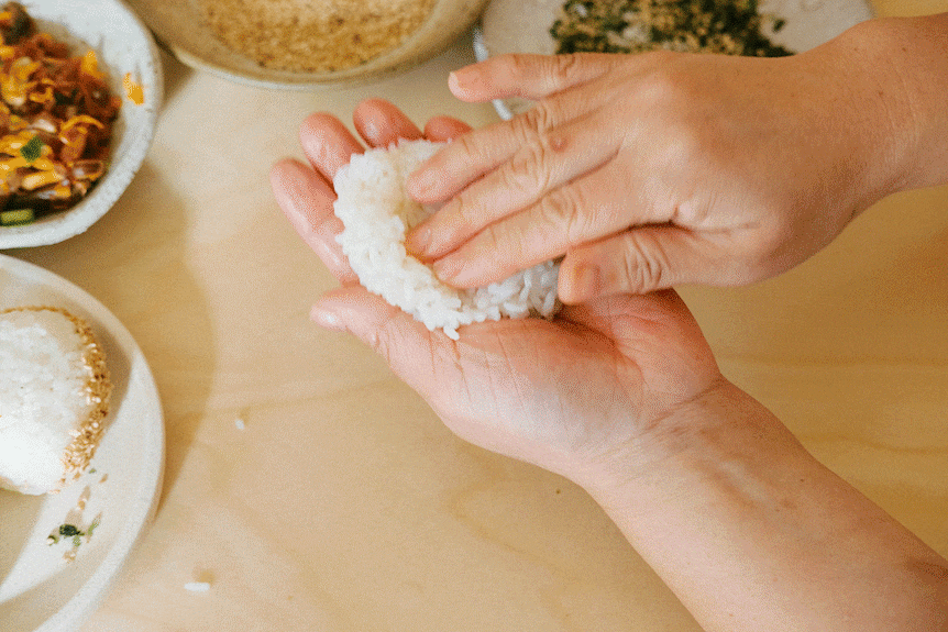 A GIF showing how to shape onigiri with your hands, from adding a filling, shaping to a triangle, rolling in sesame seeds. 