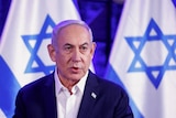 Benjamin Netanyahu stands in front of two Israeli flags. 