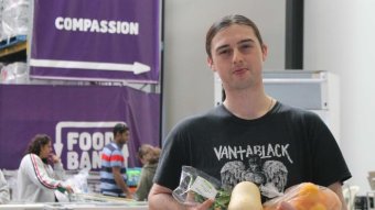 A man wearing a black t-shirt has an armful of food in front of Foodbank and compassion sign.