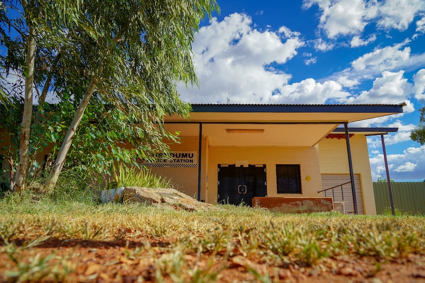 A small, single-level yellow building with a sign saying "Yuendumu Police Station", bordered by trees, grass and rocks.