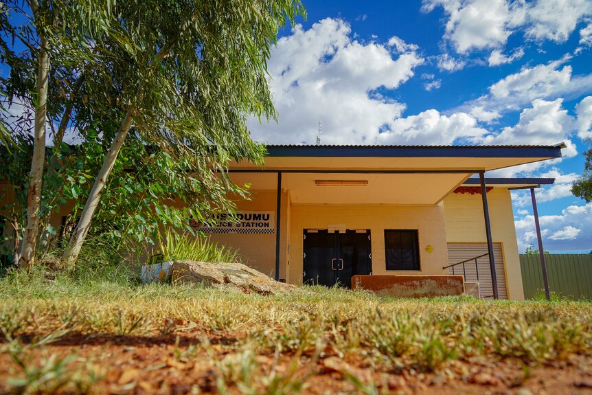 A small, single-level yellow building with a sign saying "Yuendumu Police Station", bordered by trees, grass and rocks.