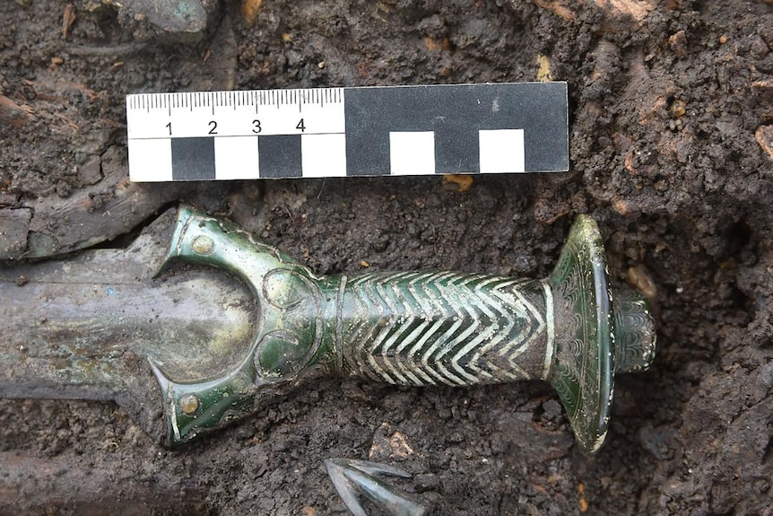 Image of a sword hilt with wave lines pictured next to a ruler in the dirt.