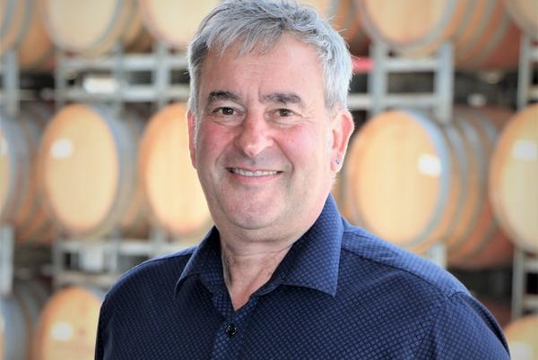 A smiling, grey-haired man stands in front of a wall racked with wine casks.